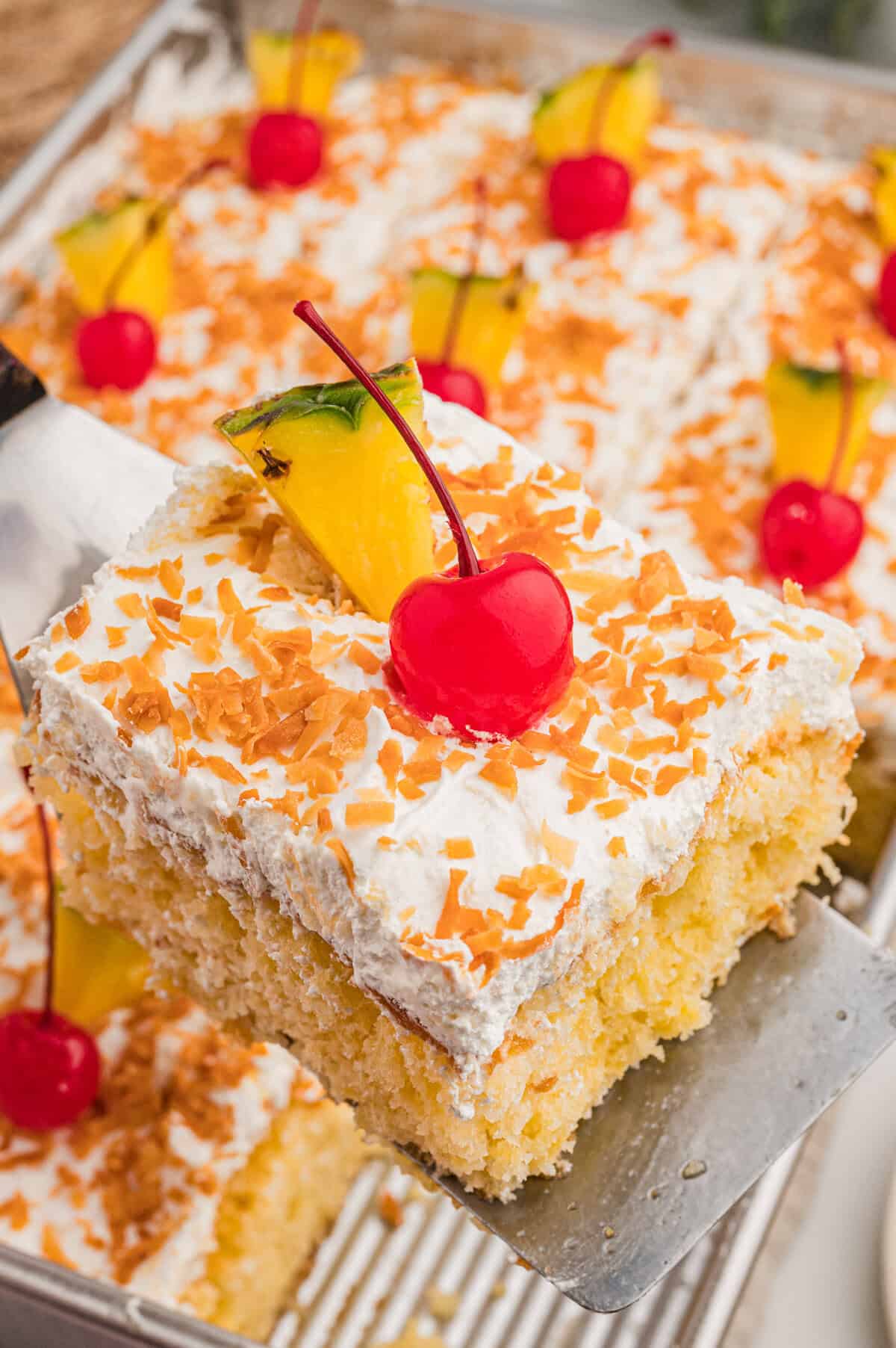 A slice of poke cake lifted on a metal server, showing yellow cake, whipped topping, toasted coconut, and a cherry.