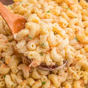 A wooden spoon lifting a scoop of creamy Hawaiian mac salad with shredded carrots and green onion from a bowl.