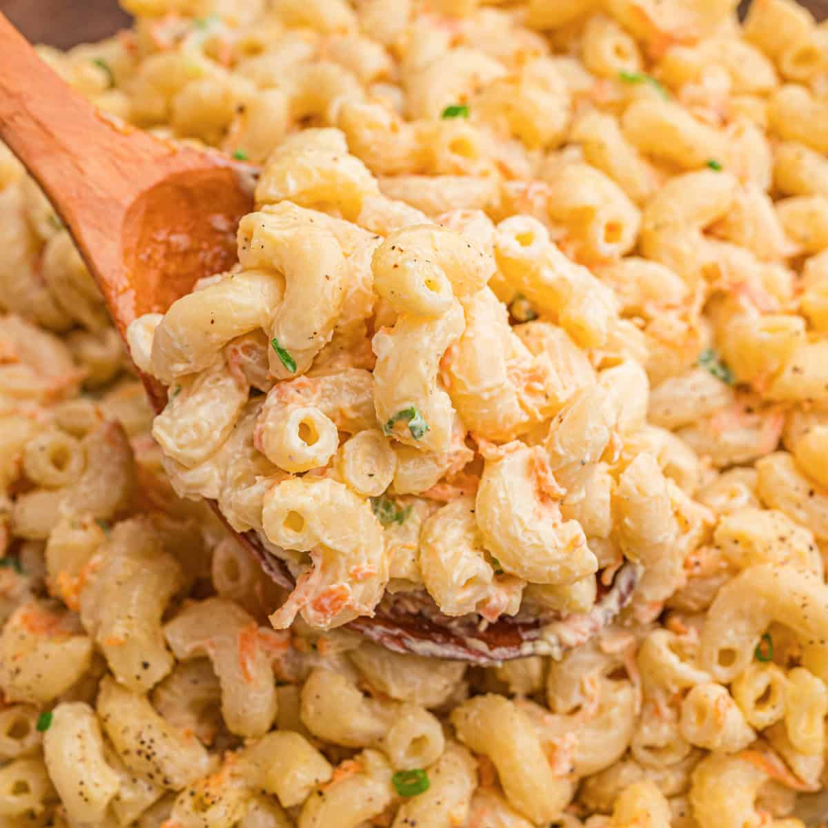 A wooden spoon lifting a scoop of creamy Hawaiian mac salad with shredded carrots and green onion from a bowl.