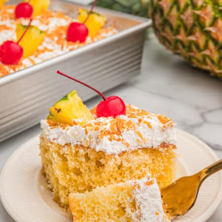 A slice of pina colada cake on a plate topped with whipped topping, toasted coconut, a pineapple wedge, and a cherry with a fresh pineapple in the background.