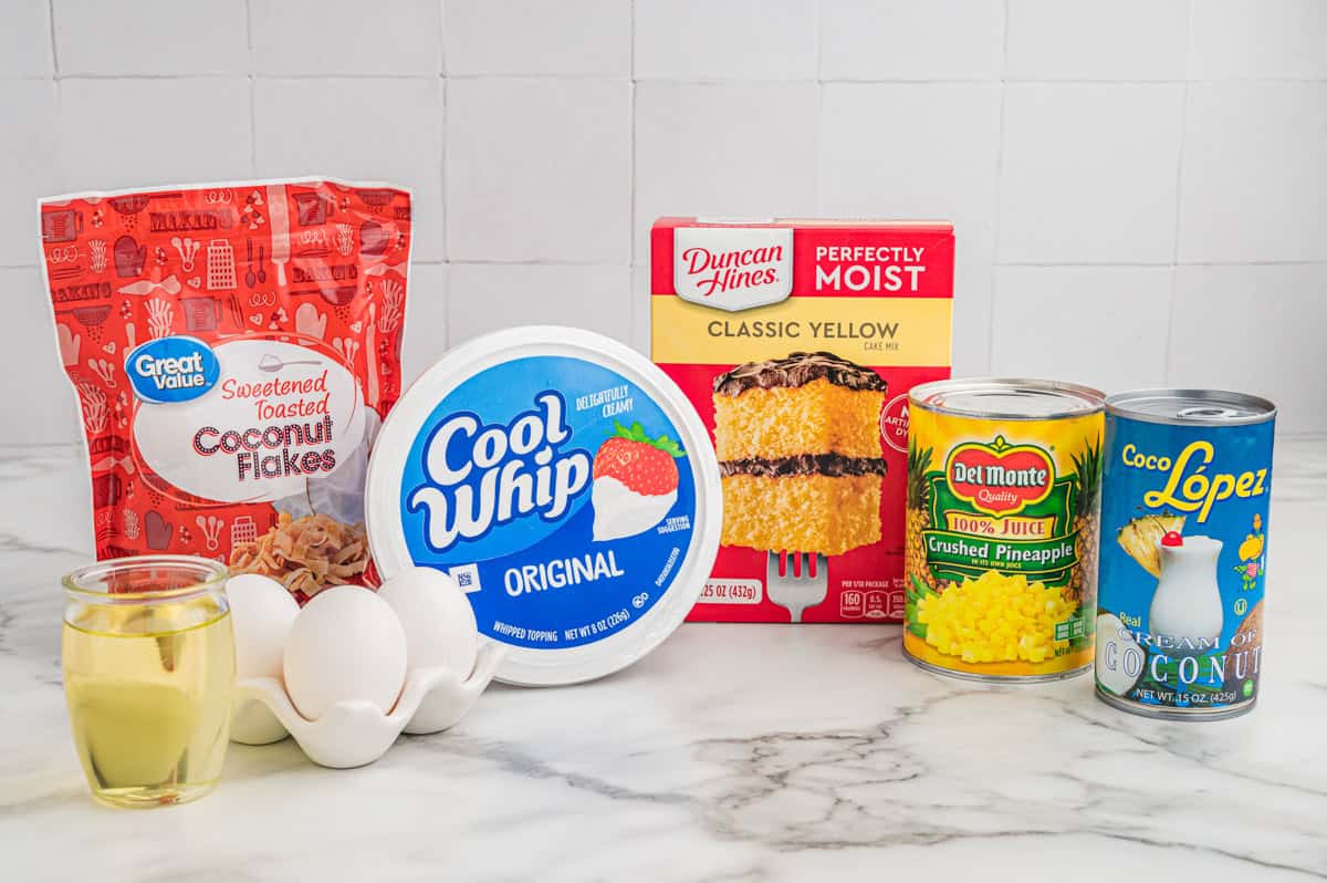 Ingredients on a marble counter: toasted coconut flakes, Cool Whip, Duncan Hines yellow cake mix, crushed pineapple, cream of coconut, eggs, and oil.