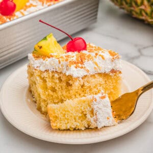 A square slice of pina colada cake on a plate with a gold fork, topped with coconut, a pineapple wedge, and a cherry.