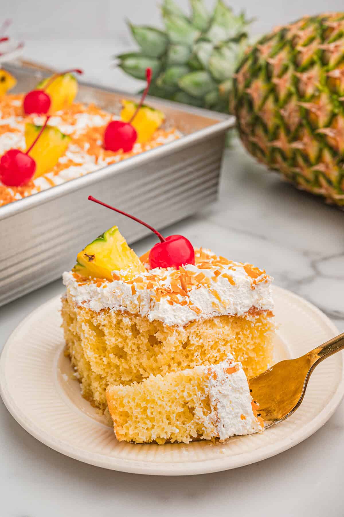 A slice of pina colada cake on a plate topped with whipped topping, toasted coconut, a pineapple wedge, and a cherry with a fresh pineapple in the background.