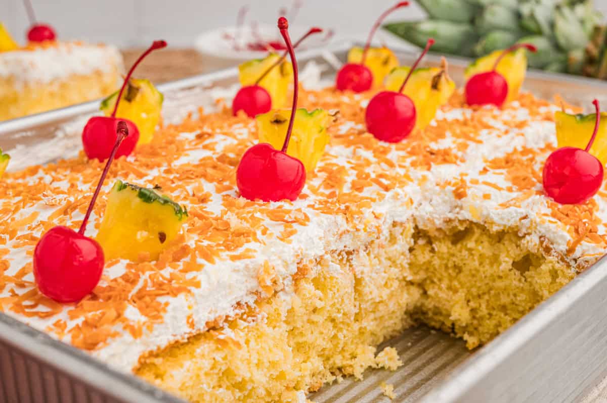 Close-up of the cake in a metal pan, cut into squares and topped with toasted coconut, maraschino cherries, and pineapple wedges.