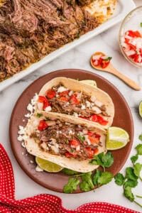 Two carne asada tacos on a brown plate topped with pico de gallo, cotija cheese, and cilantro with lime wedges.