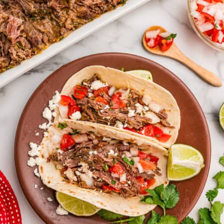 Two carne asada tacos on a brown plate topped with pico de gallo, cotija cheese, and cilantro with lime wedges.