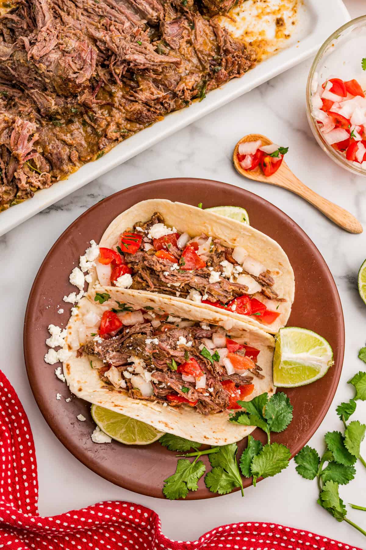 Two carne asada tacos on a brown plate topped with pico de gallo, cotija cheese, and cilantro with lime wedges.