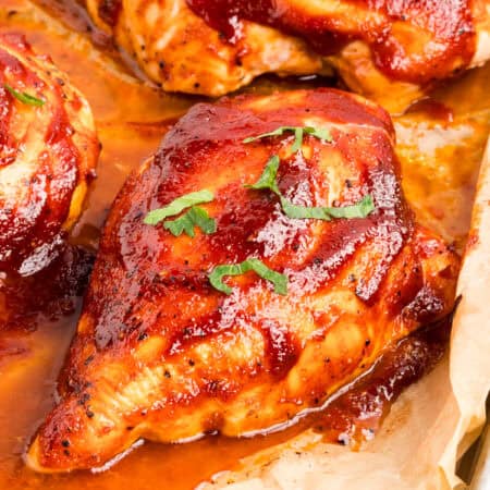 Angled close-up of a glazed barbecue chicken breast topped with parsley on a parchment-lined sheet pan.