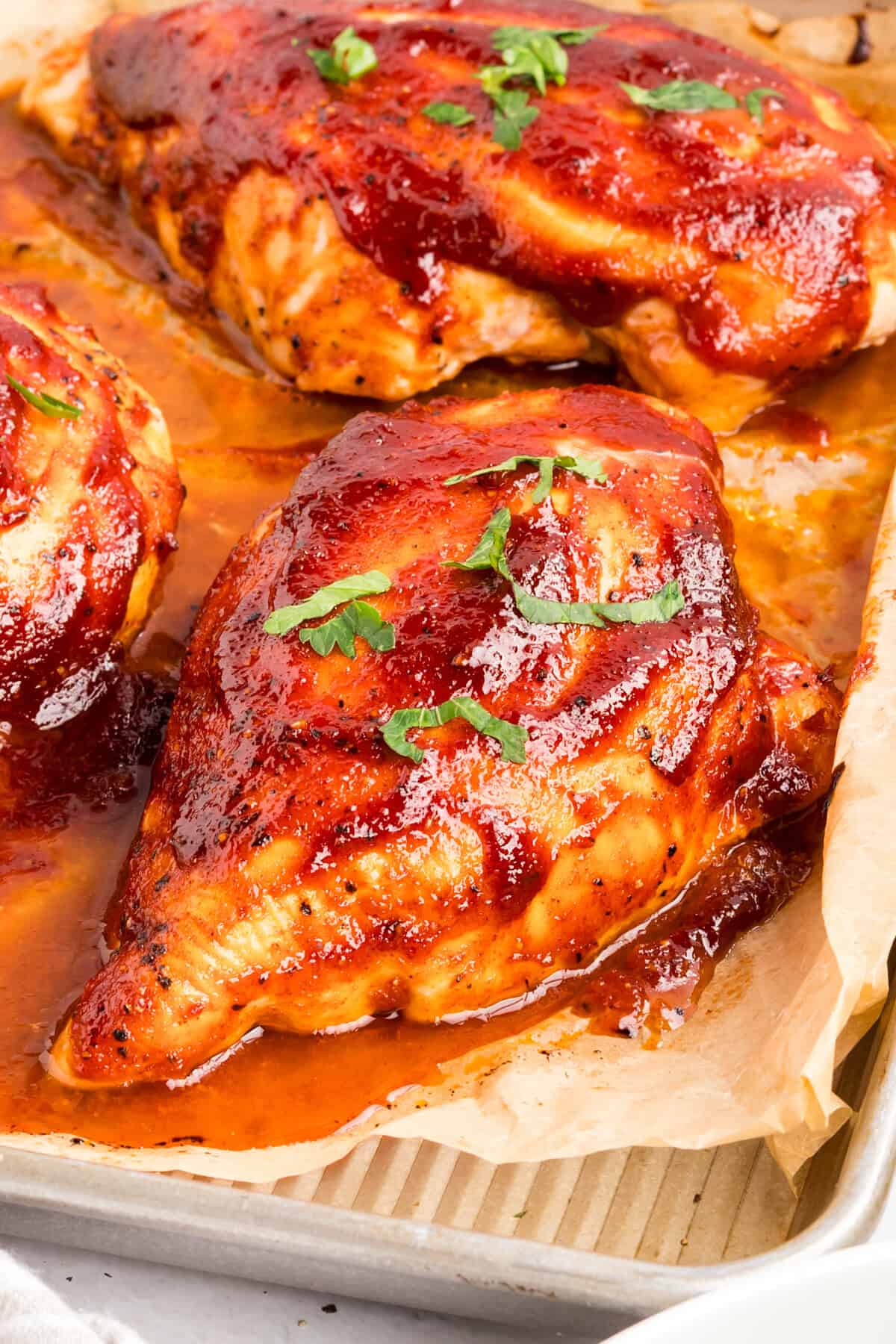 Angled close-up of a glazed barbecue chicken breast topped with parsley on a parchment-lined sheet pan.