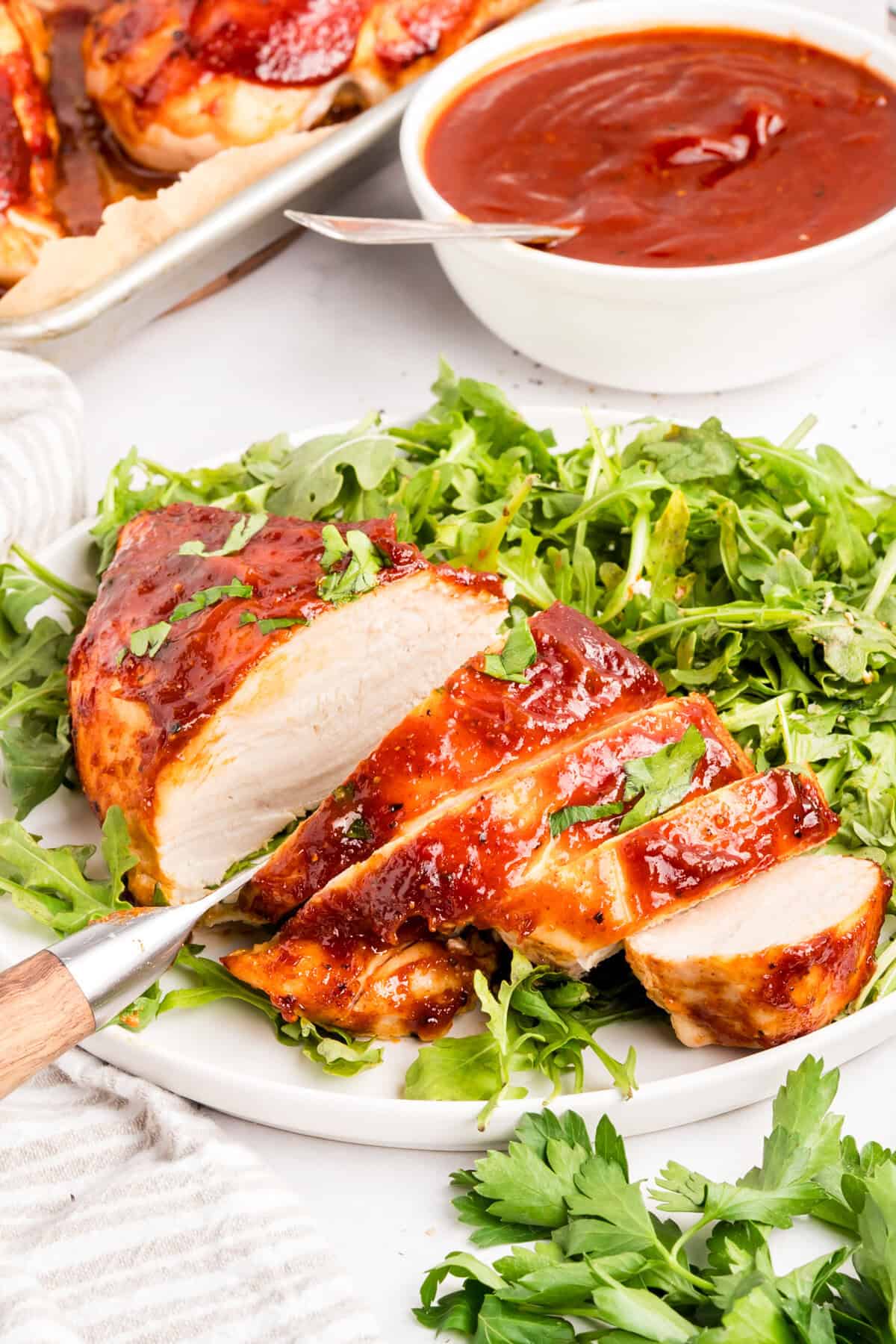 Sliced baked BBQ chicken breast on a bed of arugula with a knife and a bowl of sauce behind it.