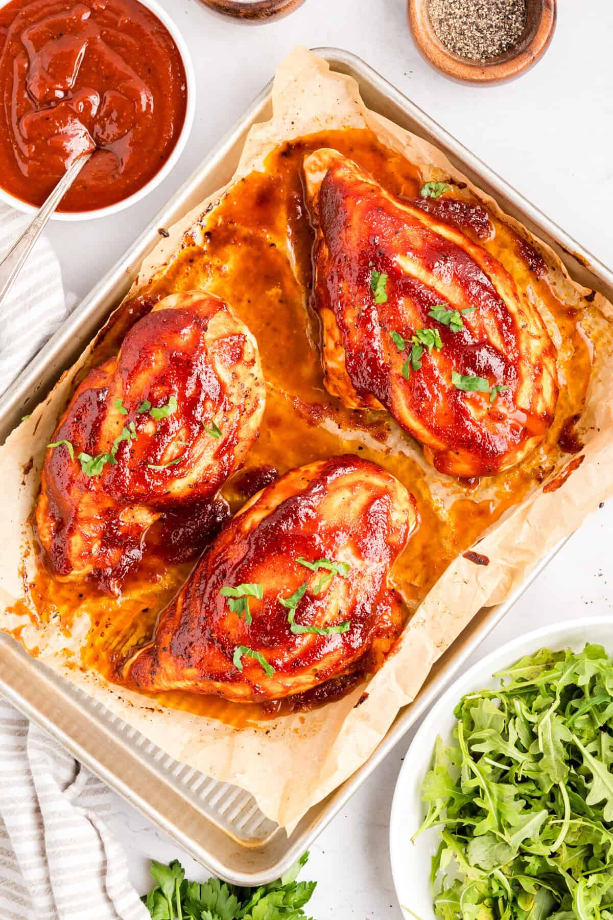 Overhead of baked BBQ chicken breast on a parchment-lined sheet pan with a bowl of BBQ sauce.