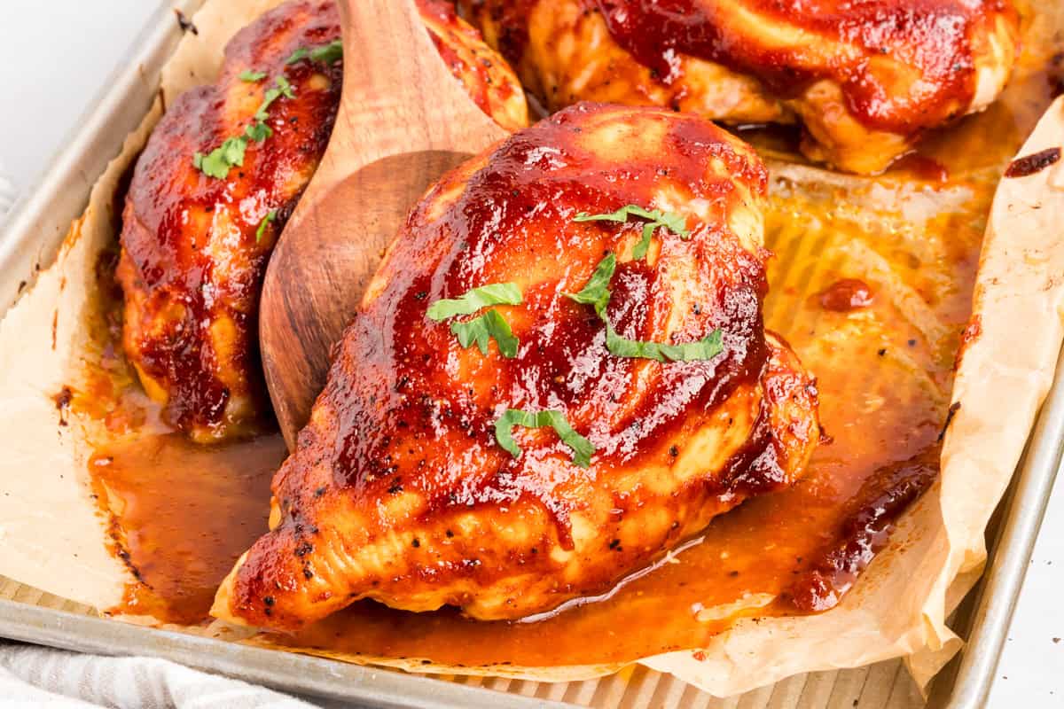 Barbecue chicken breasts with a wooden spoon tucked beside them on parchment in a metal sheet pan.