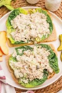Two open-faced tuna salad sandwiches on lettuce and toast with apple slices and pickles nearby.