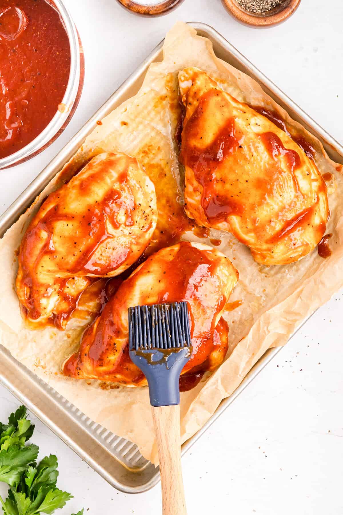 A pastry brush spreading barbecue sauce over three raw chicken breasts on a parchment-lined sheet pan.