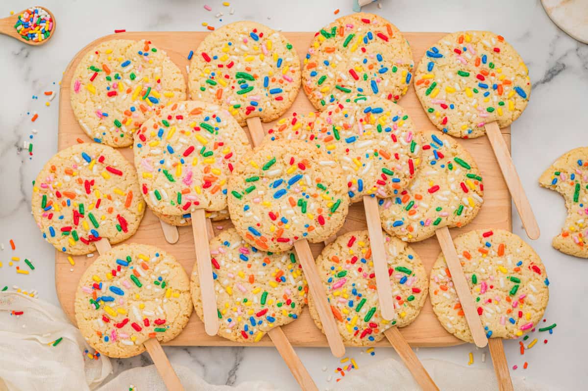 Sugar cookies on sticks topped with rainbow sprinkles arranged on a wooden cutting board.