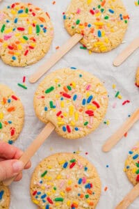 Cookies on a stick topped with rainbow sprinkles, one lifted by hand from a parchment-lined surface of matching cookies.