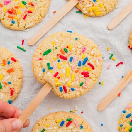 Cookies on a stick topped with rainbow sprinkles, one lifted by hand from a parchment-lined surface of matching cookies.