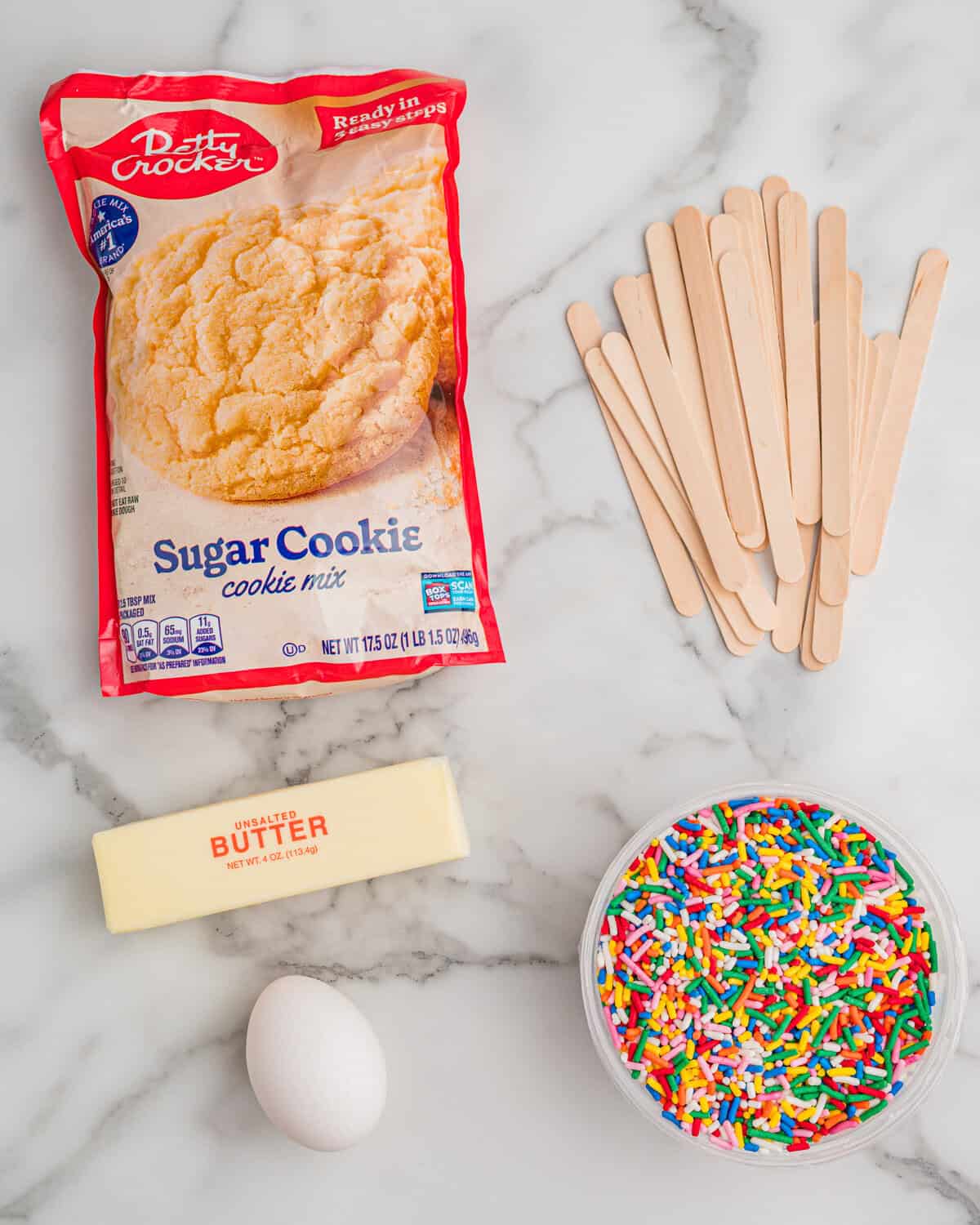 Ingredients on a marble counter: Betty Crocker sugar cookie mix, wooden popsicle sticks, a stick of unsalted butter, an egg, and a bowl of rainbow sprinkles.