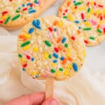 Cookies on a stick topped with rainbow sprinkles, one held by hand over a cream napkin with two more behind.