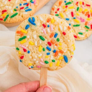 Cookies on a stick topped with rainbow sprinkles, one held by hand over a cream napkin with two more behind.