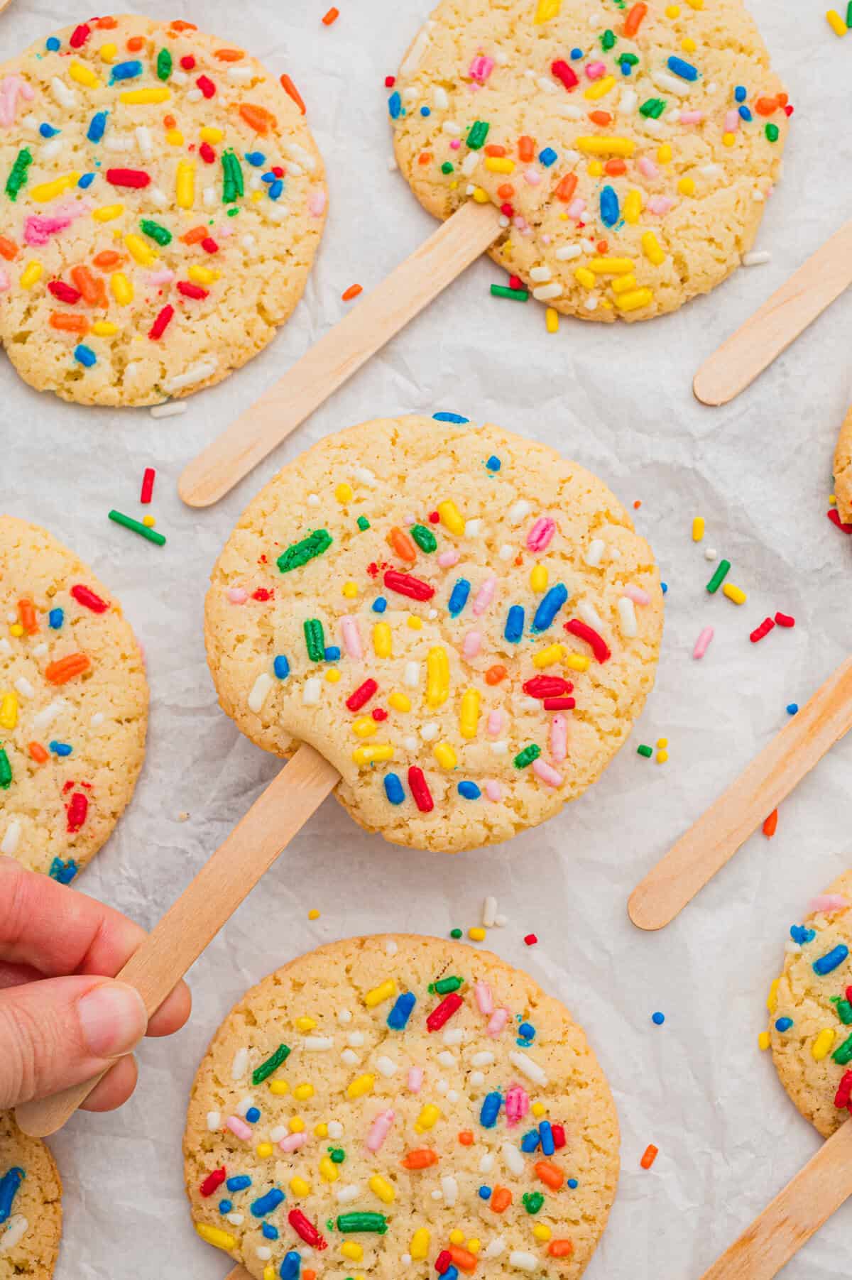 Cookies on a stick topped with rainbow sprinkles, one lifted by hand from a parchment-lined surface of matching cookies.