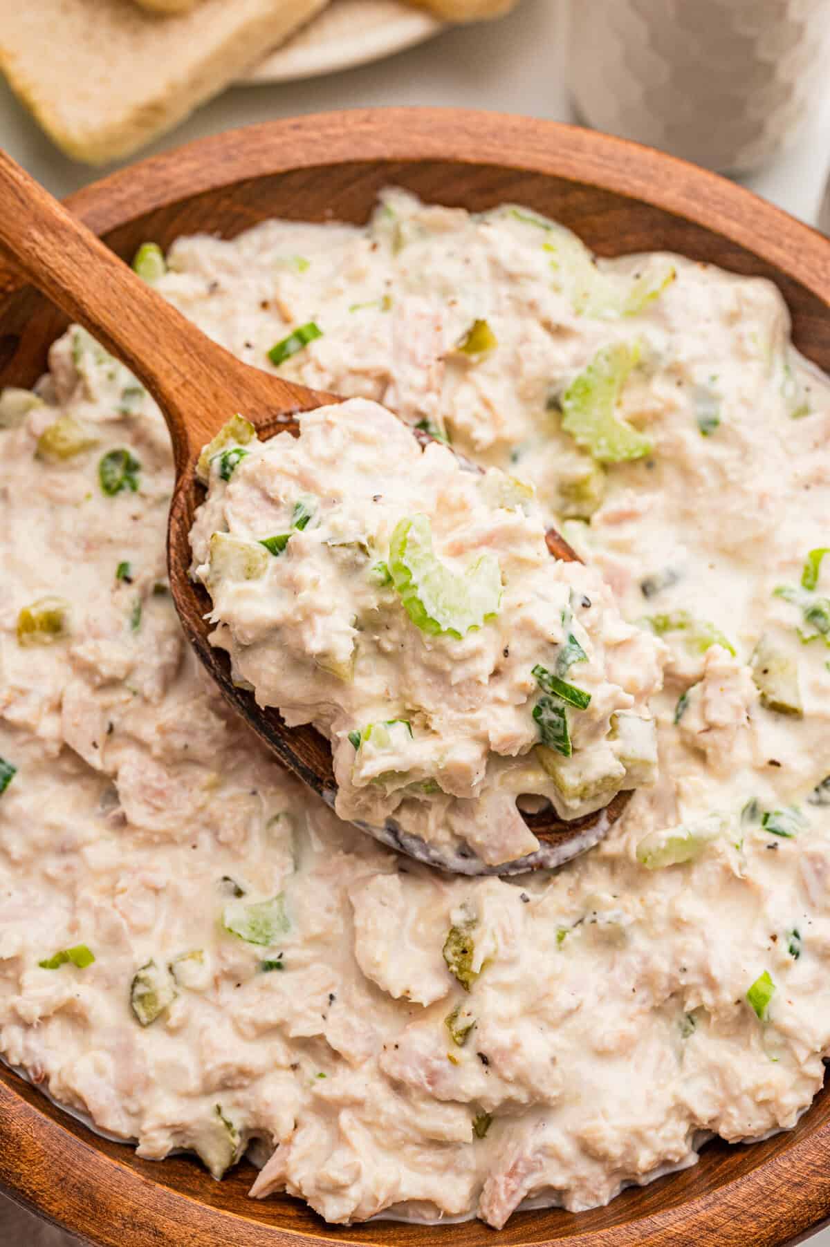 A wooden spoon lifting creamy tuna salad from a wooden bowl flecked with celery and pepper.
