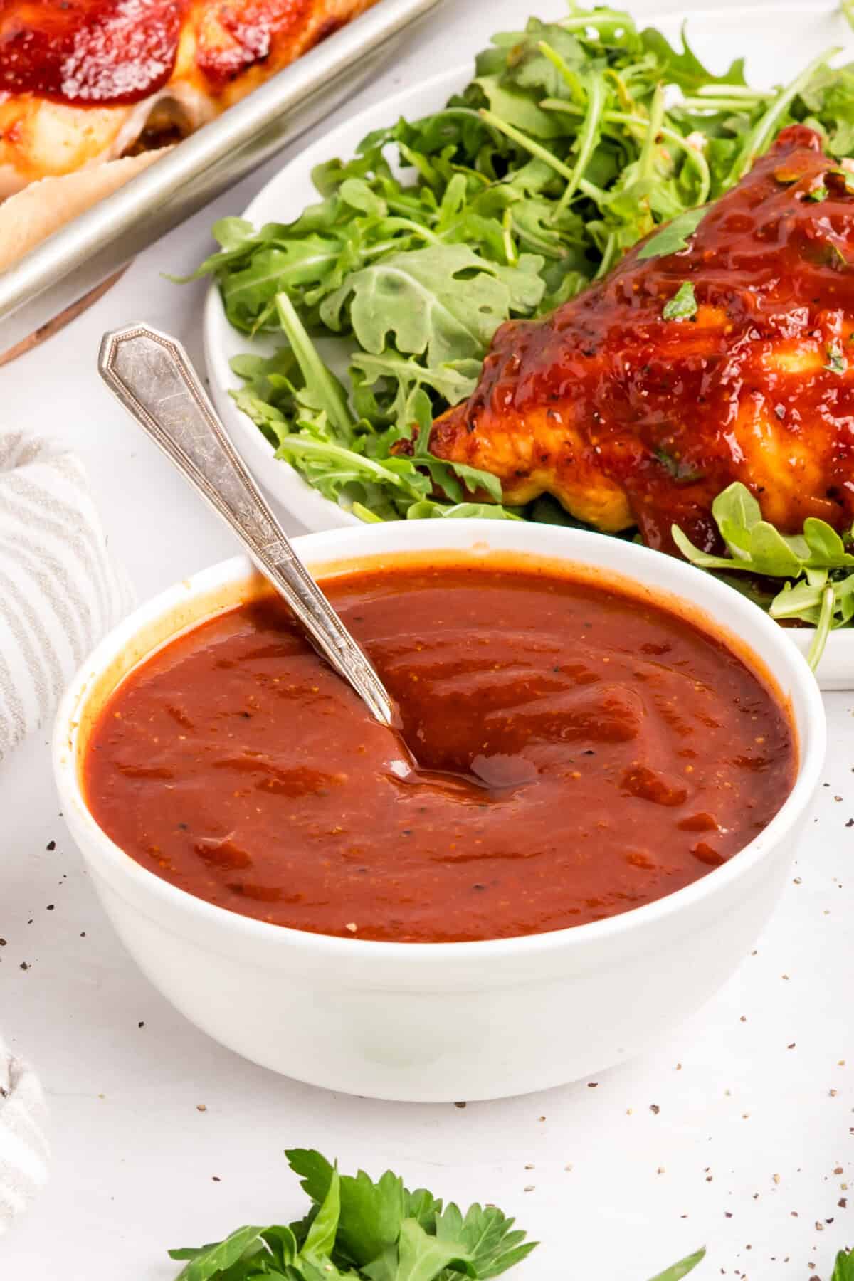 Homemade BBQ sauce in a white bowl with a spoon, a plate of BBQ chicken over arugula behind it.