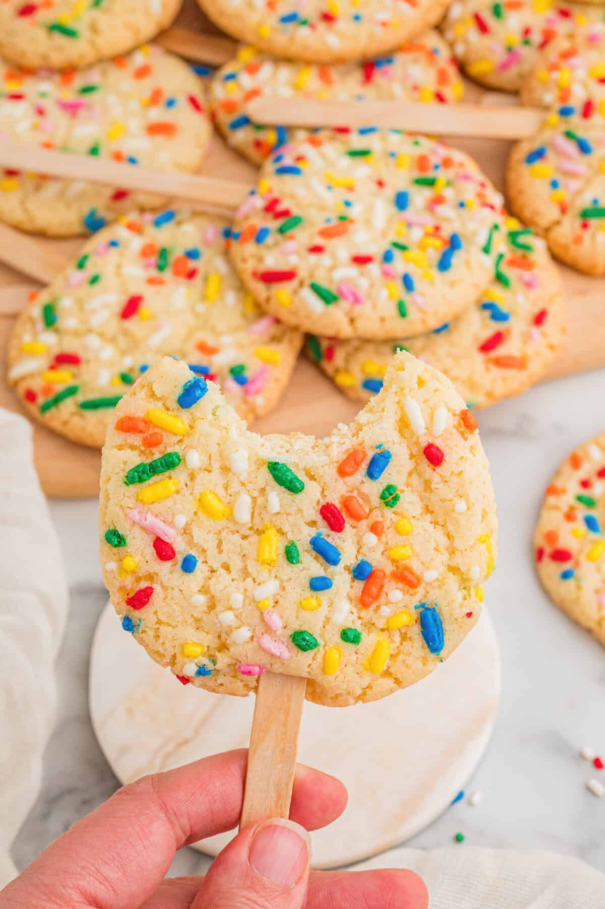 A hand holding a sprinkle sugar cookie on a stick with a bite taken out, more cookies on a wooden board behind.