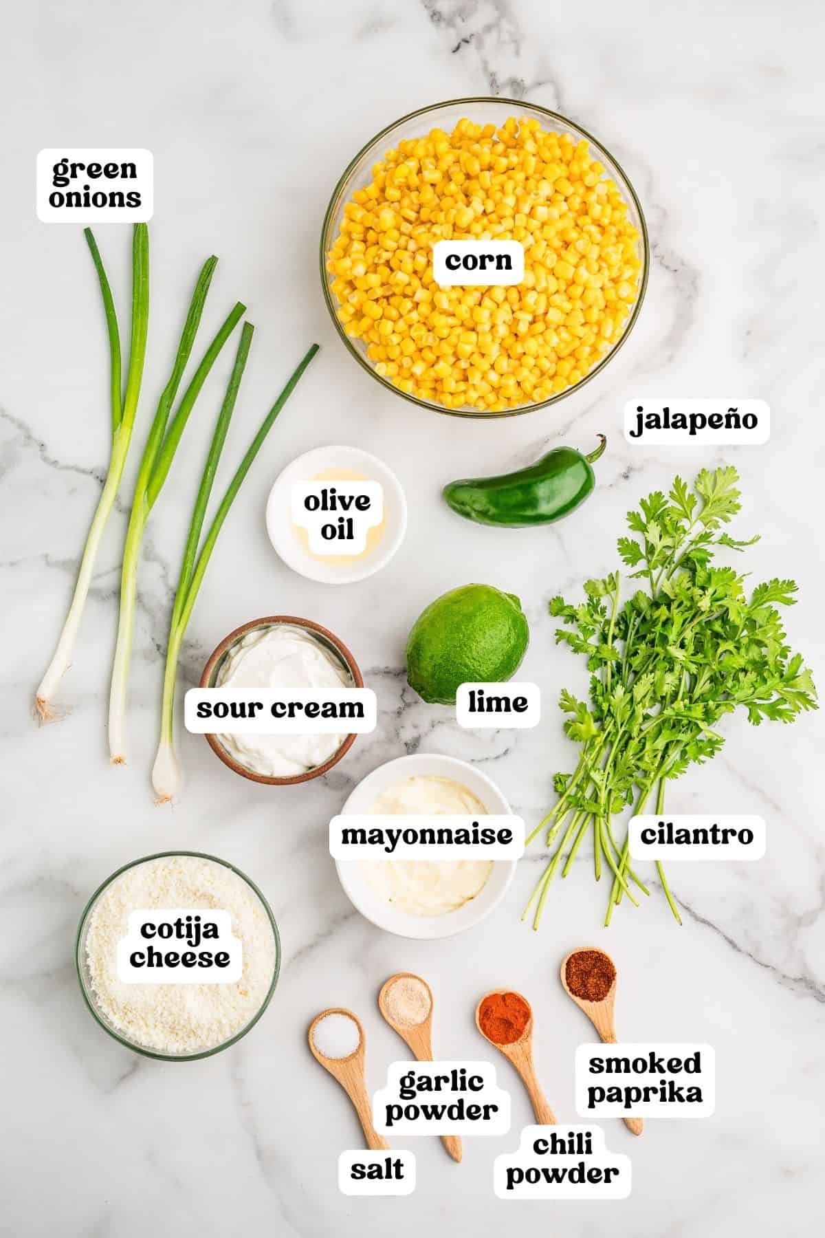 Labeled ingredients on marble: corn, green onions, jalapeno, olive oil, sour cream, lime, cilantro, mayonnaise, cotija cheese, and spices.