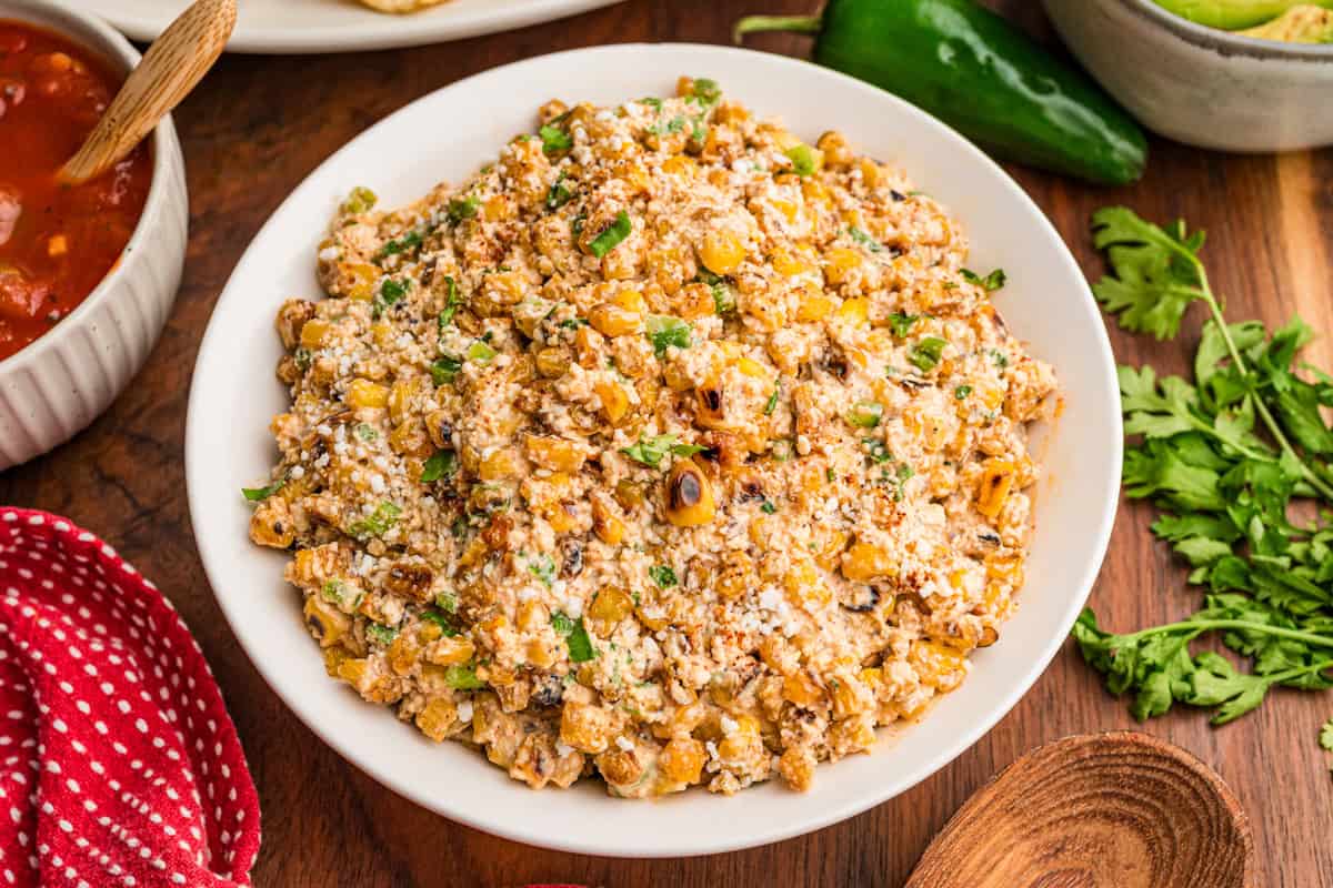 Mexican street corn dip in a white bowl with charred corn, cotija, and cilantro, tortilla chips and salsa nearby.