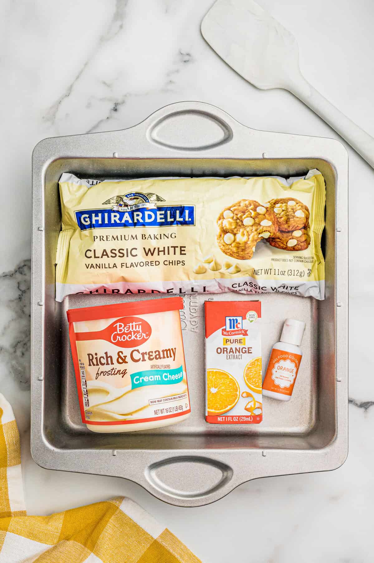 Ingredients in a metal pan: Ghirardelli white chocolate chips, cream cheese frosting, orange extract, and orange food coloring.