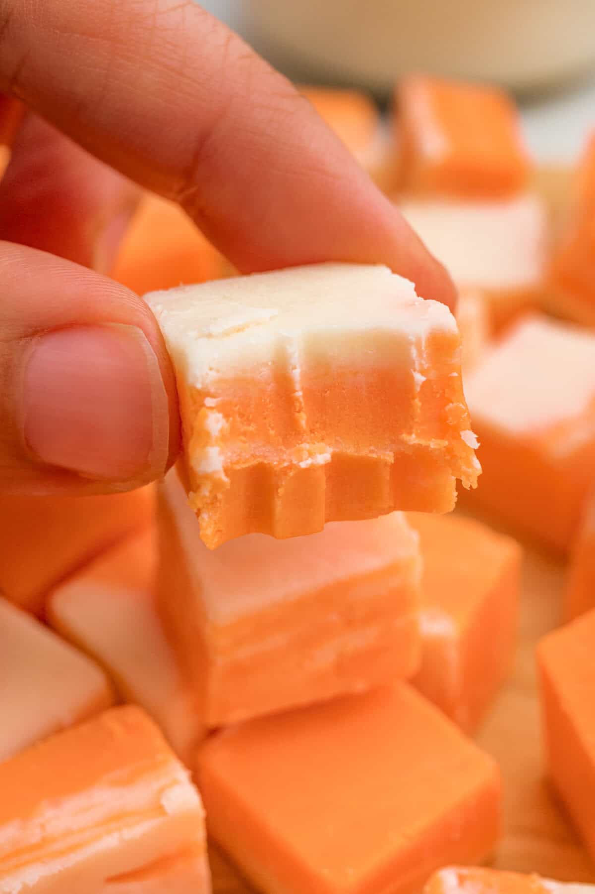A hand holding a square of orange and white fudge with a bite taken out.
