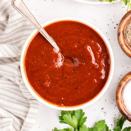 Overhead bowl of homemade barbecue sauce with a spoon, a plate of sauced chicken and small dishes of salt and pepper alongside.