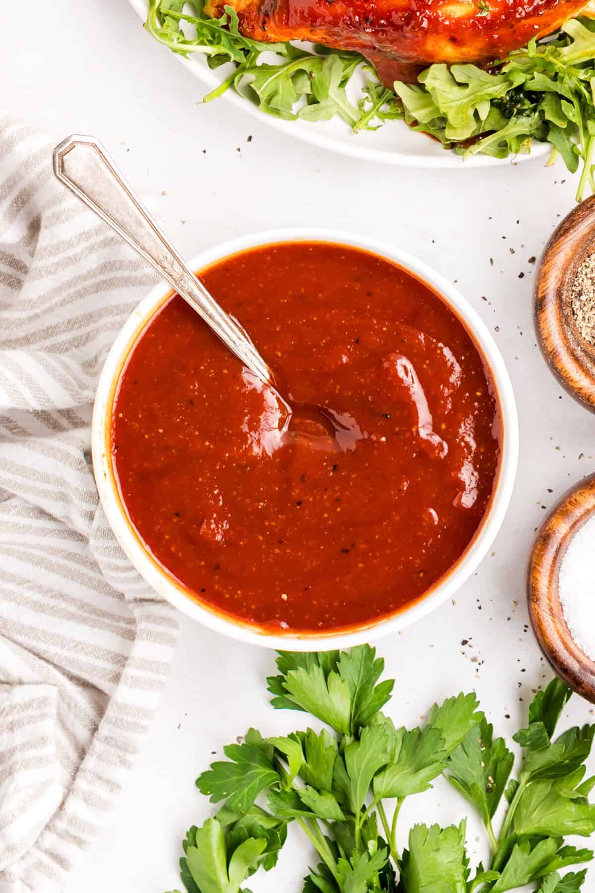 Overhead bowl of homemade barbecue sauce with a spoon, a plate of sauced chicken and small dishes of salt and pepper alongside.