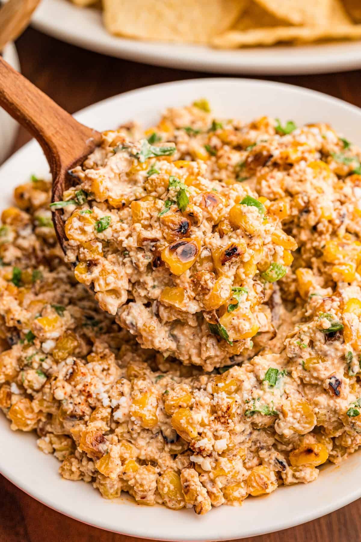 A wooden spoon lifting a scoop of creamy corn dip from a white bowl, tortilla chips in the background.