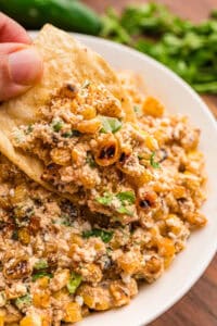 A hand dipping a tortilla chip into corn dip loaded with charred corn, cotija, and cilantro.