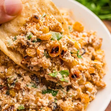 A hand dipping a tortilla chip into corn dip loaded with charred corn, cotija, and cilantro.