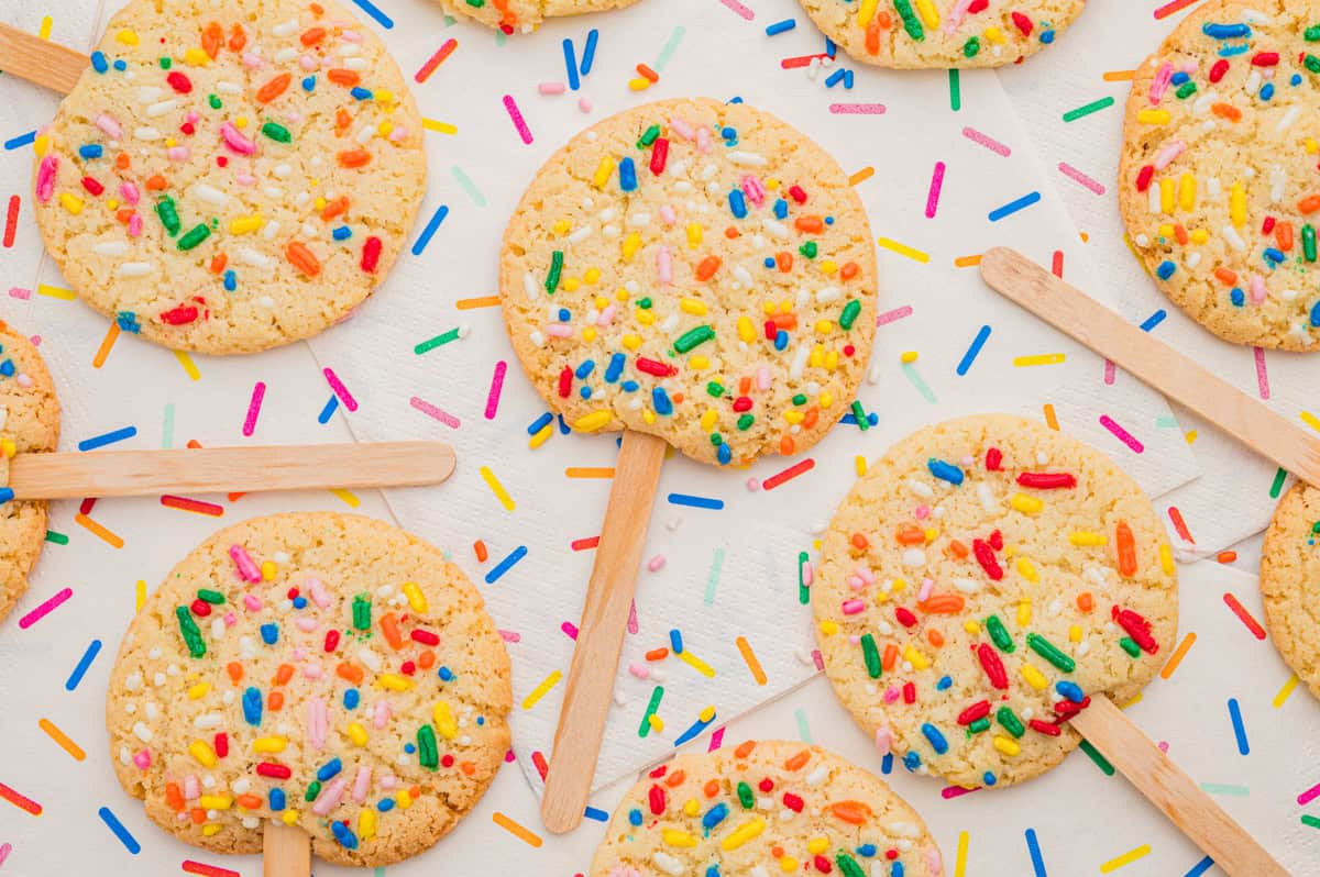 Rows of sprinkle sugar cookies on sticks on a napkin scattered with colorful sprinkles.