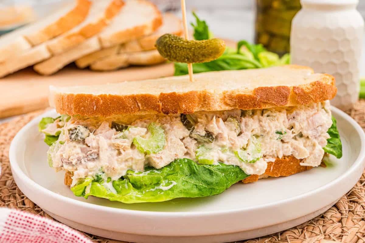 A tuna salad sandwich on white bread with lettuce and a baby dill pickle on a pick.