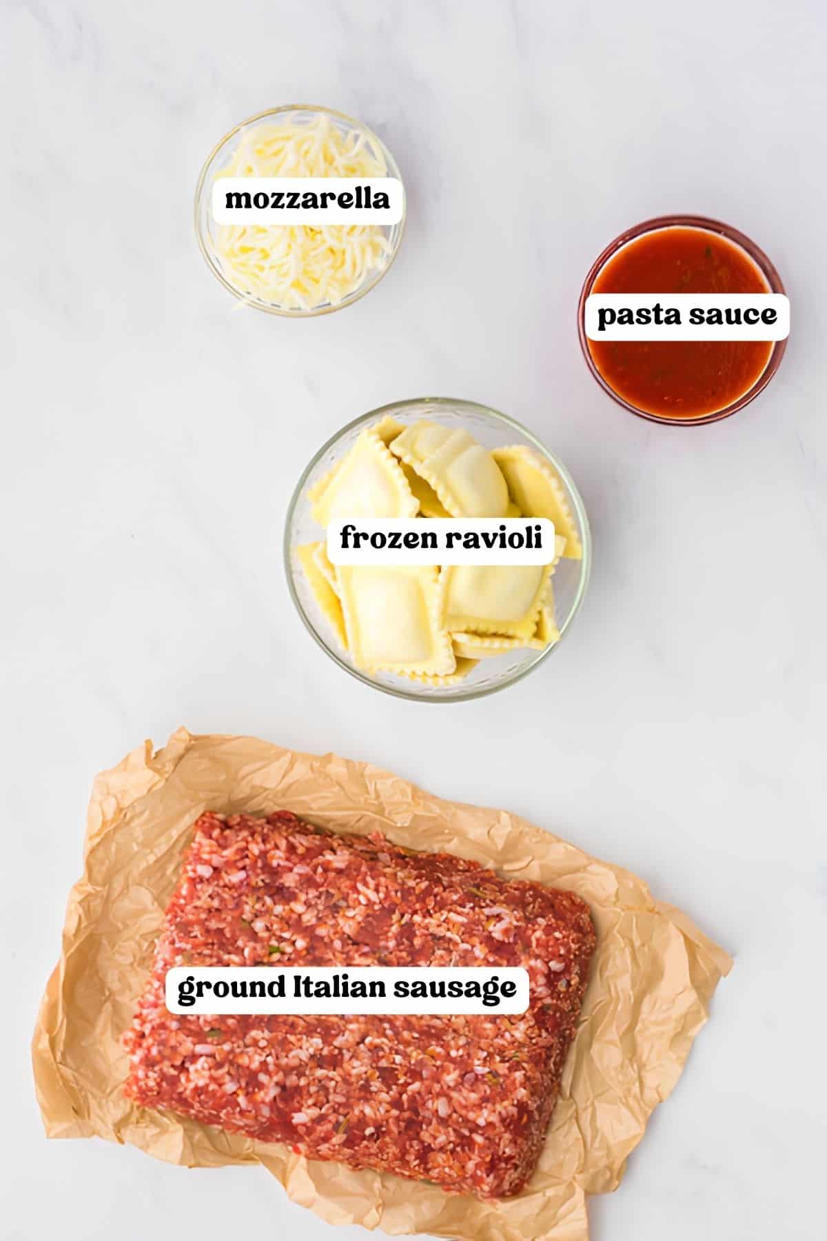 Shredded mozzarella, jarred pasta sauce, frozen ravioli, and ground Italian sausage arranged in bowls on a marble counter.