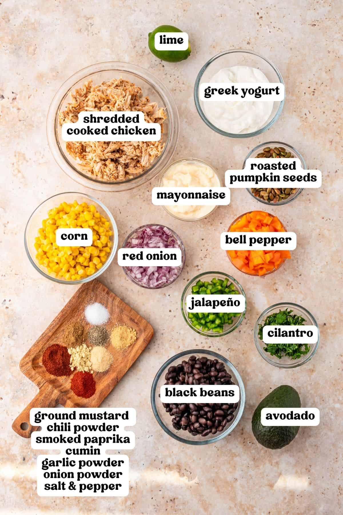 Shredded cooked chicken, Greek yogurt, mayonnaise, corn, black beans, bell pepper, red onion, jalapeno, cilantro, pumpkin seeds, avocado, lime, and spices in bowls.