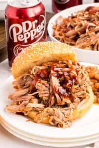A barbecue pulled pork sandwich on a sesame bun with sauce, baked beans and a Dr Pepper can beside it.