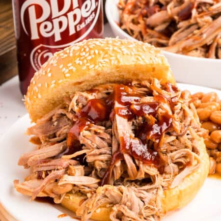 A barbecue pulled pork sandwich on a sesame bun with sauce, baked beans and a Dr Pepper can beside it.