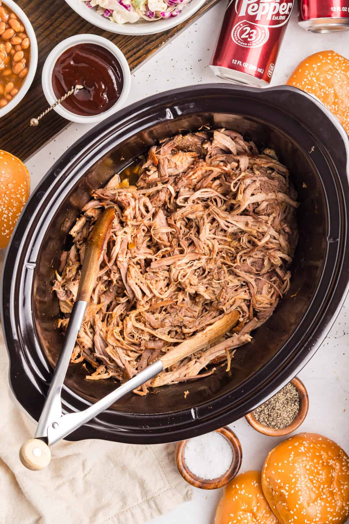 Overhead of shredded pork in a black slow cooker with wooden tongs, surrounded by buns, baked beans, coleslaw, and Dr Pepper.