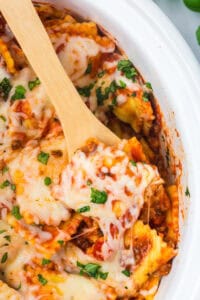 A wooden spoon lifting a cheesy serving of ravioli lasagna from the slow cooker.