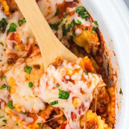 A wooden spoon lifting a cheesy serving of ravioli lasagna from the slow cooker.