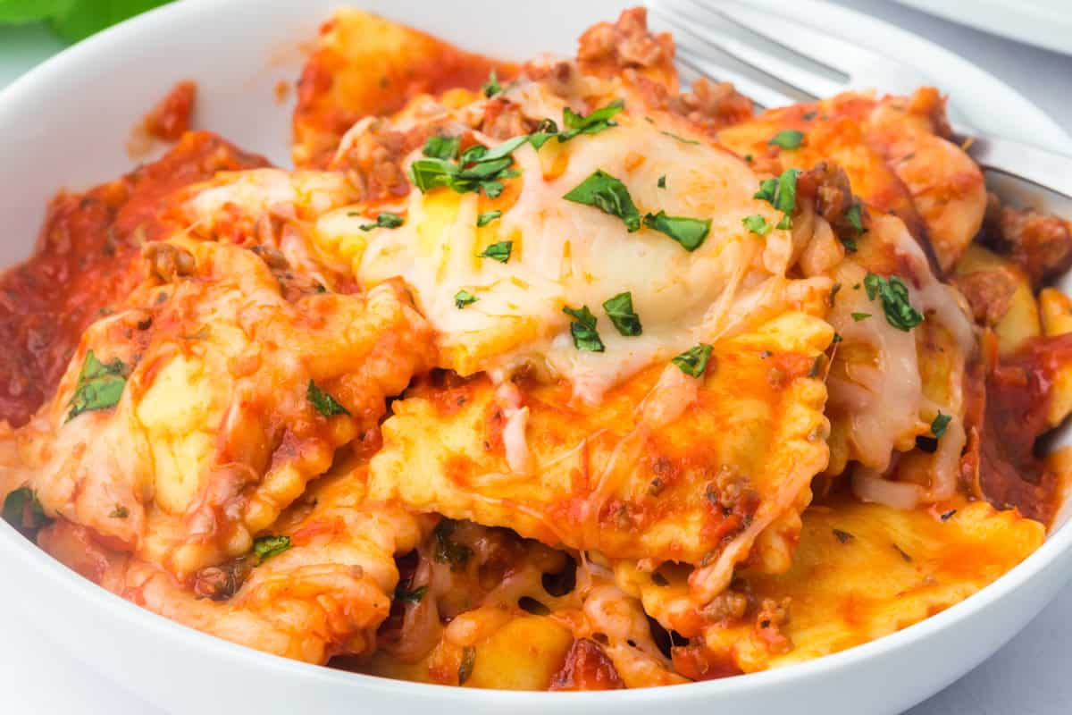 Close-up of crockpot ravioli lasagna in a white bowl with sausage, red sauce, melted cheese, and basil.