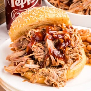 A barbecue pulled pork sandwich on a sesame bun with sauce, baked beans and a Dr Pepper can beside it.
