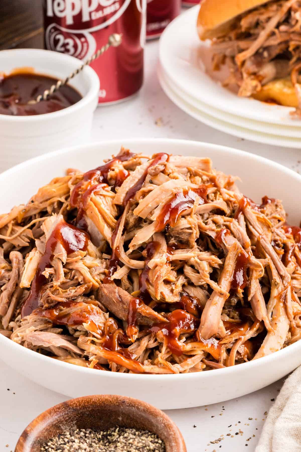 Dr Pepper pulled pork in a white bowl drizzled with barbecue sauce, a Dr Pepper can and a sandwich behind it.