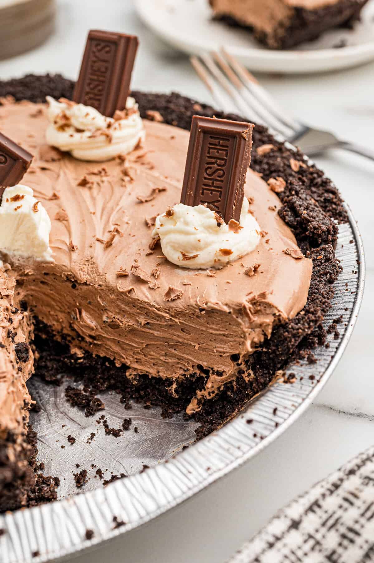 A whole chocolate pie in a foil pan with one slice removed, topped with whipped cream and Hershey's bars.